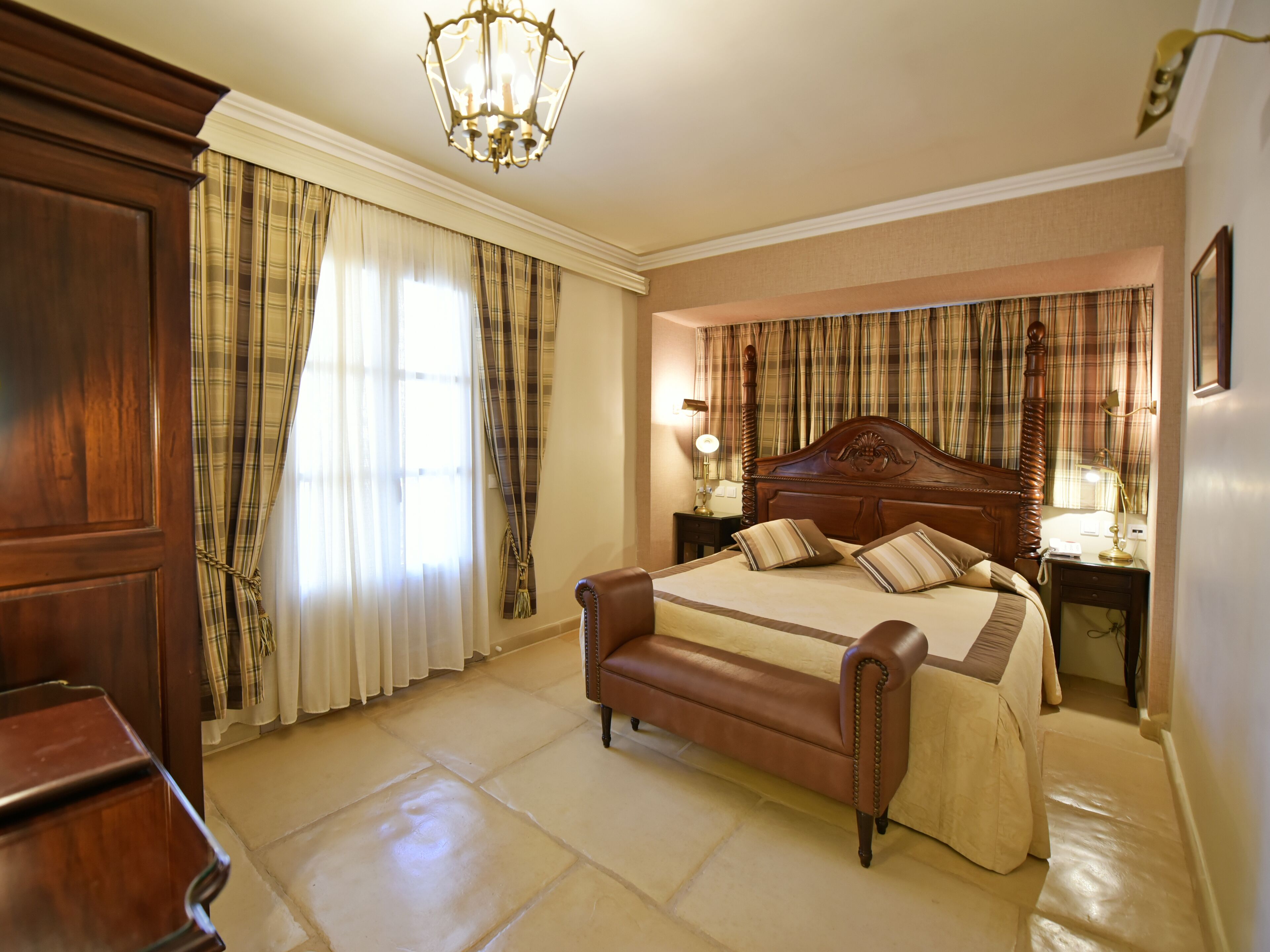 A bedroom with a large bed, wooden headboard, and curtains. There is a nightstand with a lamp and a dresser.