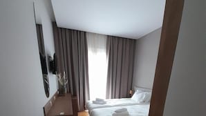 Une chambre d’hôtel moderne avec un plancher en bois, un lit recouvert de draps blancs, une petite table surmontée d’un vase et un téléviseur fixé au mur.