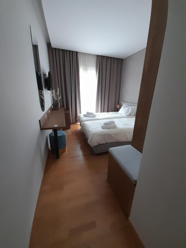 A modern hotel room with a wooden floor, a bed with white linens, a small table with a vase, and a TV mounted on the wall.