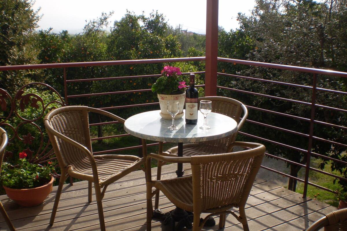 A round table with wicker chairs on a balcony, a bottle of wine, and two wine glasses.