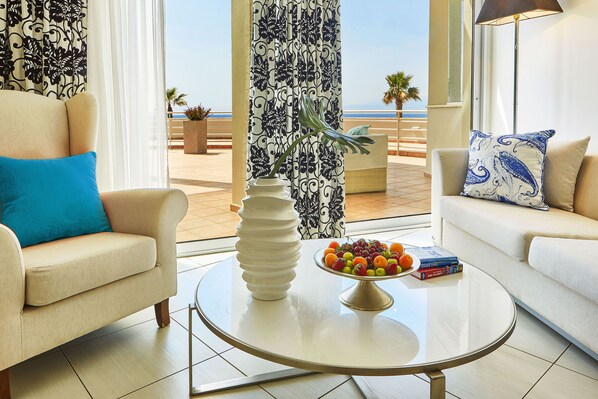 A living room with a view of the sea, a round coffee table with a fruit bowl and books, and two sofas with patterned cushions.