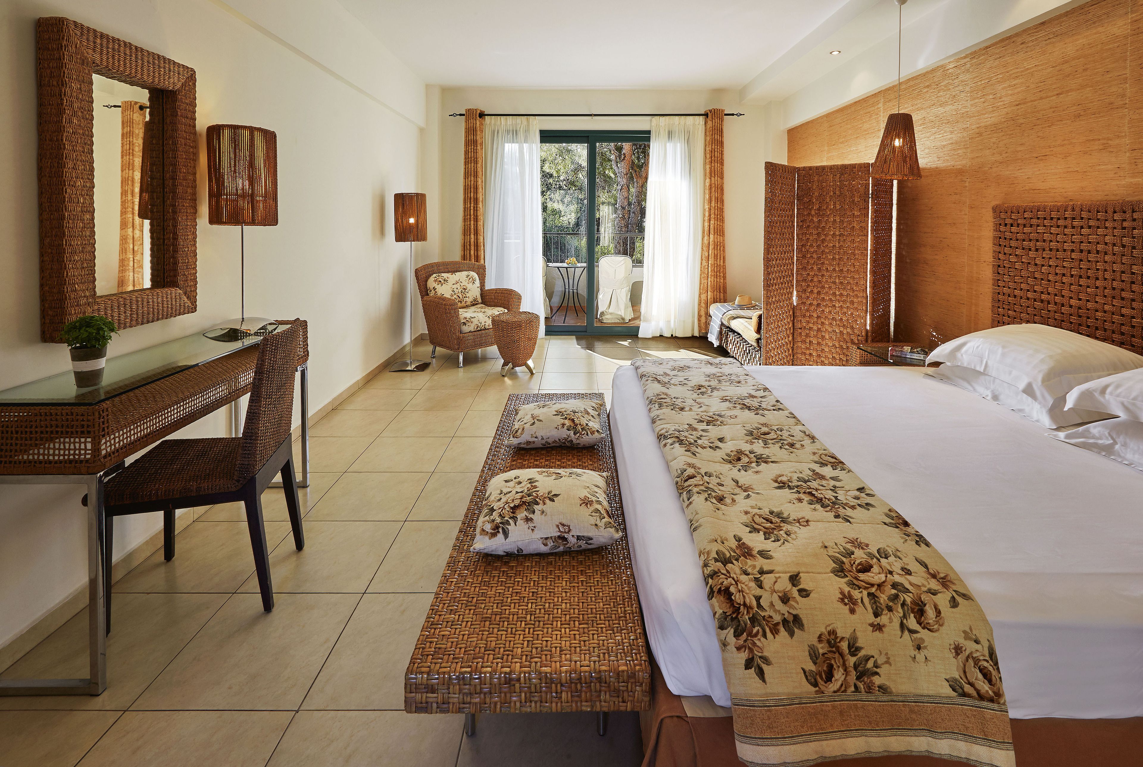Quarto de hotel com cama, escrivaninha, cadeira, banco, mesa e varanda com vista para árvores.