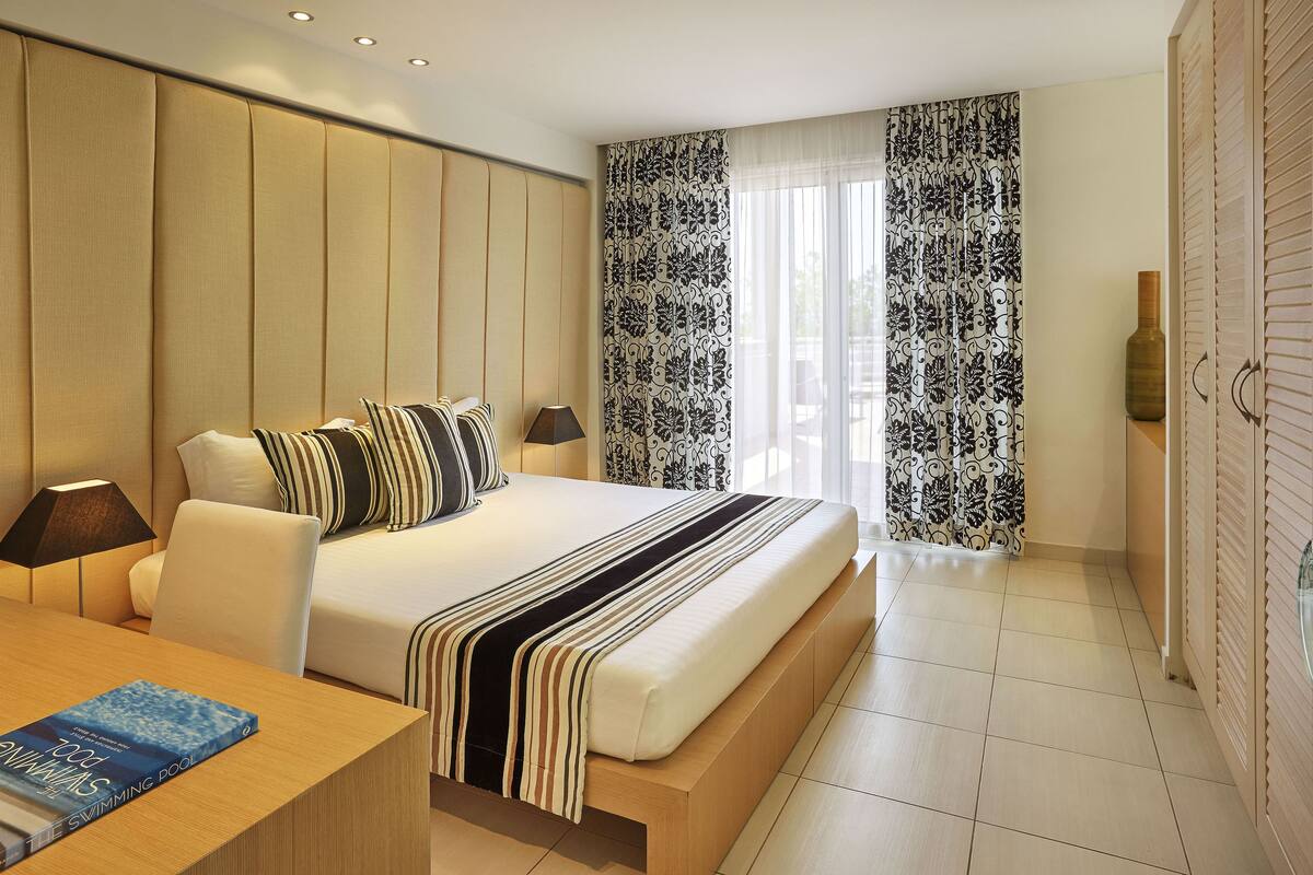 Une chambre d’hôtel moderne avec un grand lit, une table de chevet sur laquelle se trouve un livre et une lampe, et une vue sur l’extérieur à travers des rideaux à motifs.