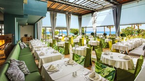 A restaurant with multiple tables and chairs, a green sofa, and a view of the ocean. - Ilio Mare Resort Hotel by GHH (Thasos)