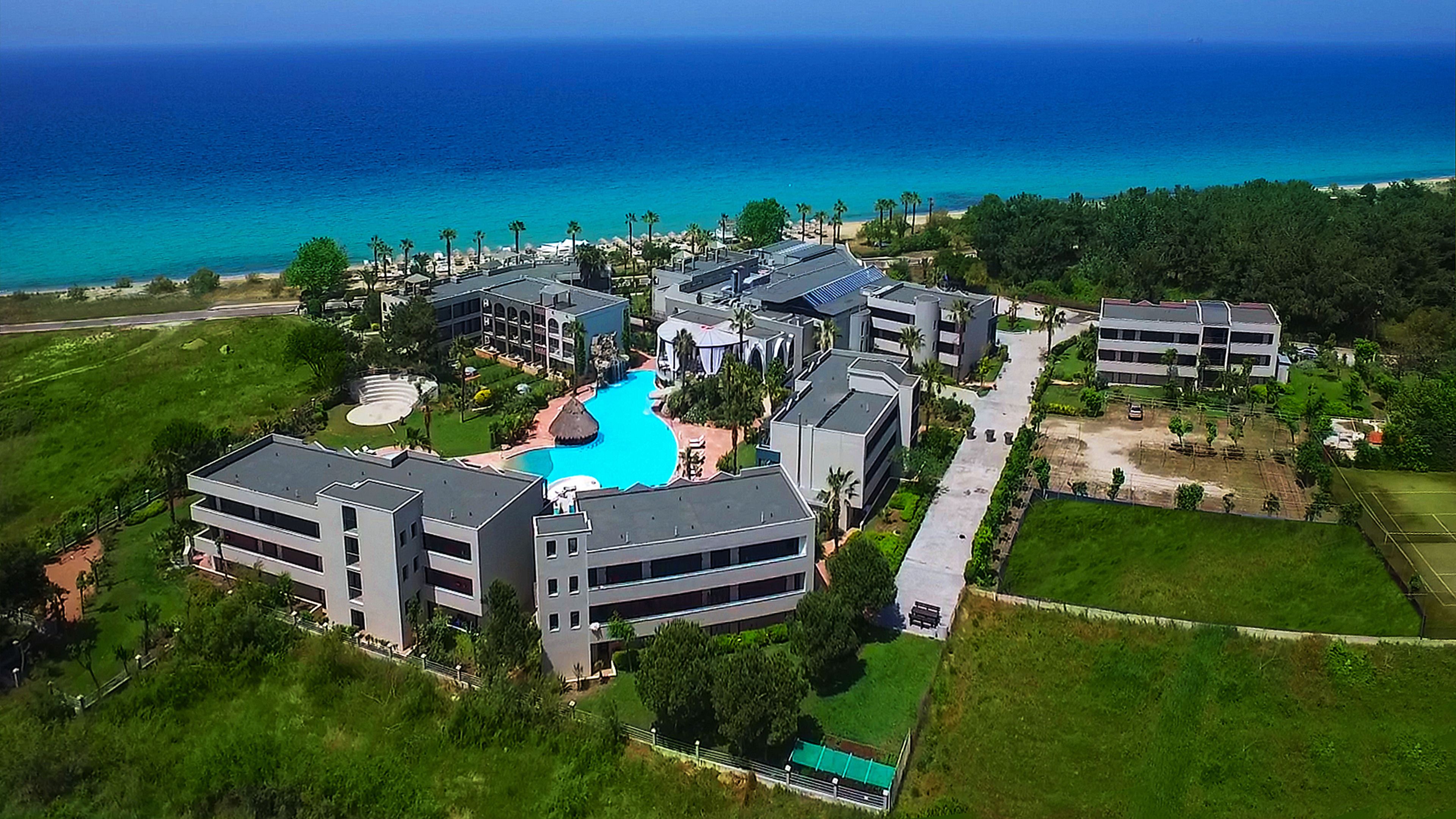 An aerial view of a resort with multiple buildings, a central swimming pool, and a view of the ocean.