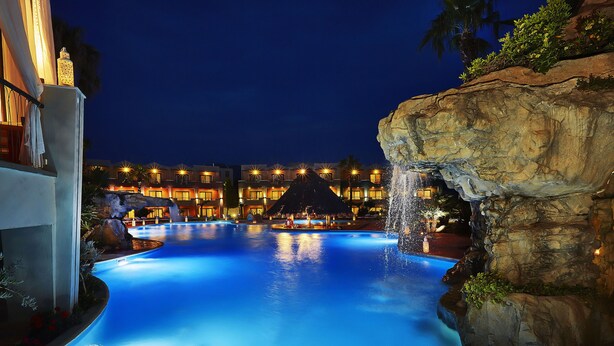 A nighttime swimming pool with a waterfall feature, surrounded by a resort building with multiple balconies and lights. - Ilio Mare Resort Hotel by GHH (Thasos)
