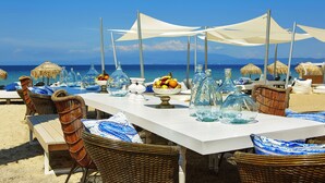 A beachside dining setup with wicker chairs, a white table, and thatched umbrellas. - Ilio Mare Resort Hotel by GHH (Thasos)
