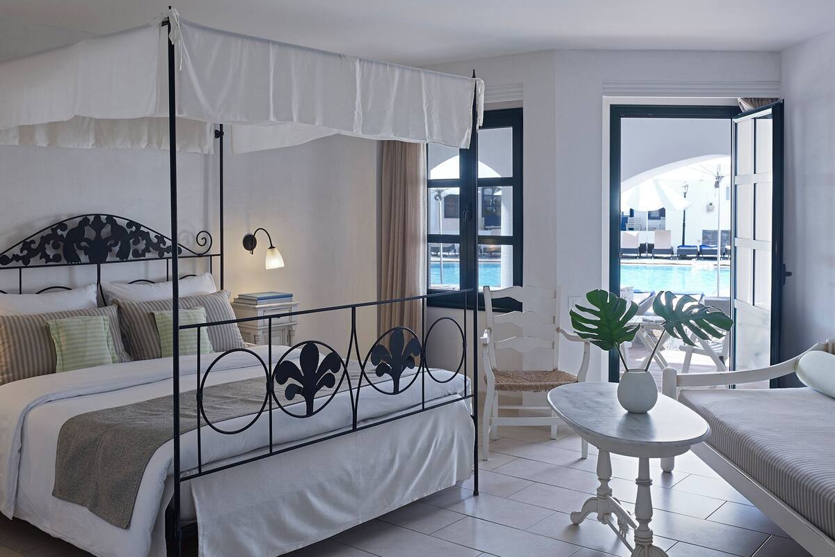 A bedroom with a four-poster bed, a bedside table with a lamp, a chair, and a view of a swimming pool through an open door.