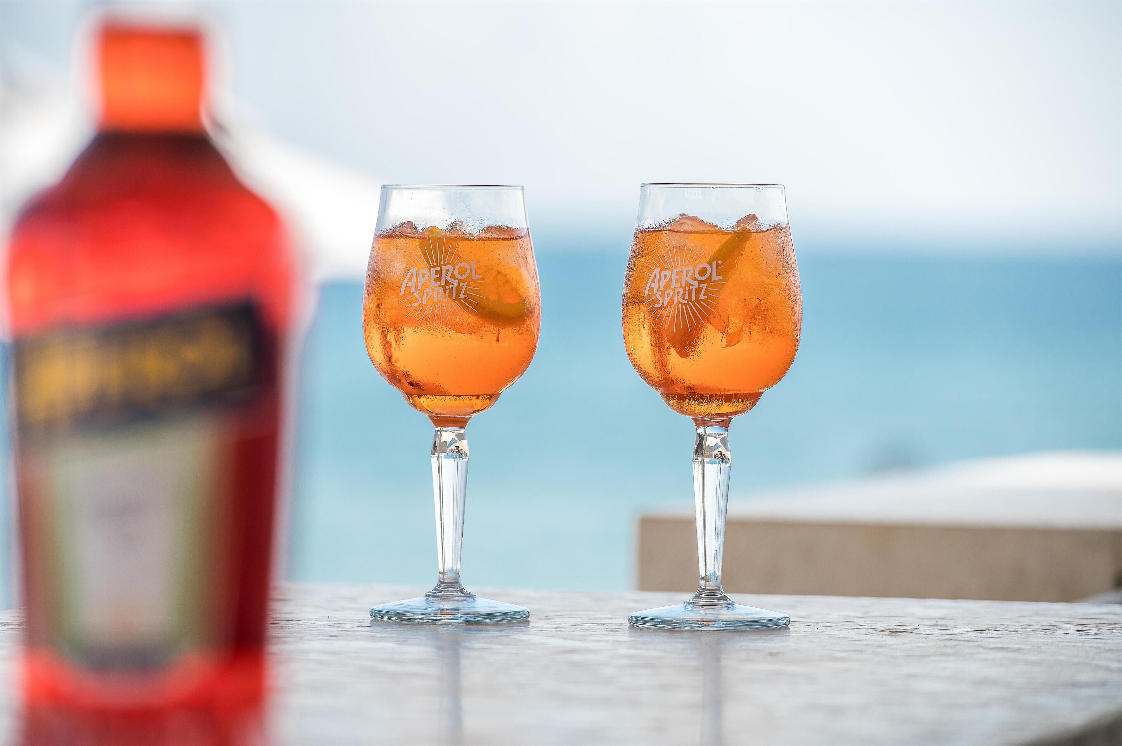 Two glasses of Aperol Spritz on a table with a blurred bottle in the background.