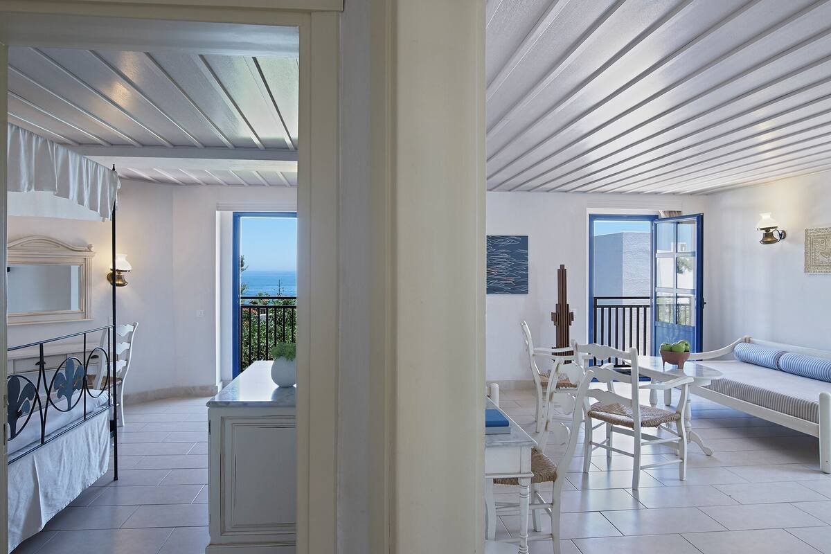 A bright, open living area with a balcony overlooking the sea, white furniture, and a tiled floor.