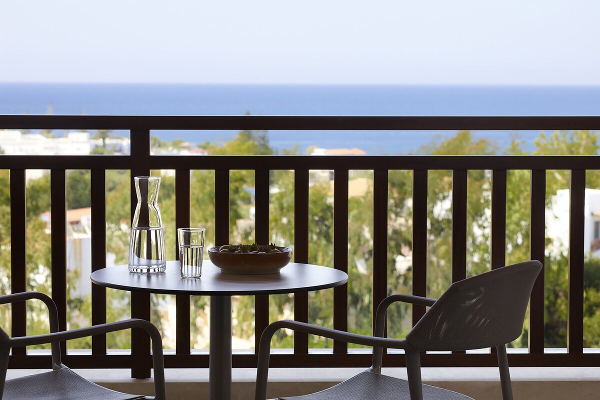 A balcony with a table and chairs, overlooking a view of the sea and greenery.