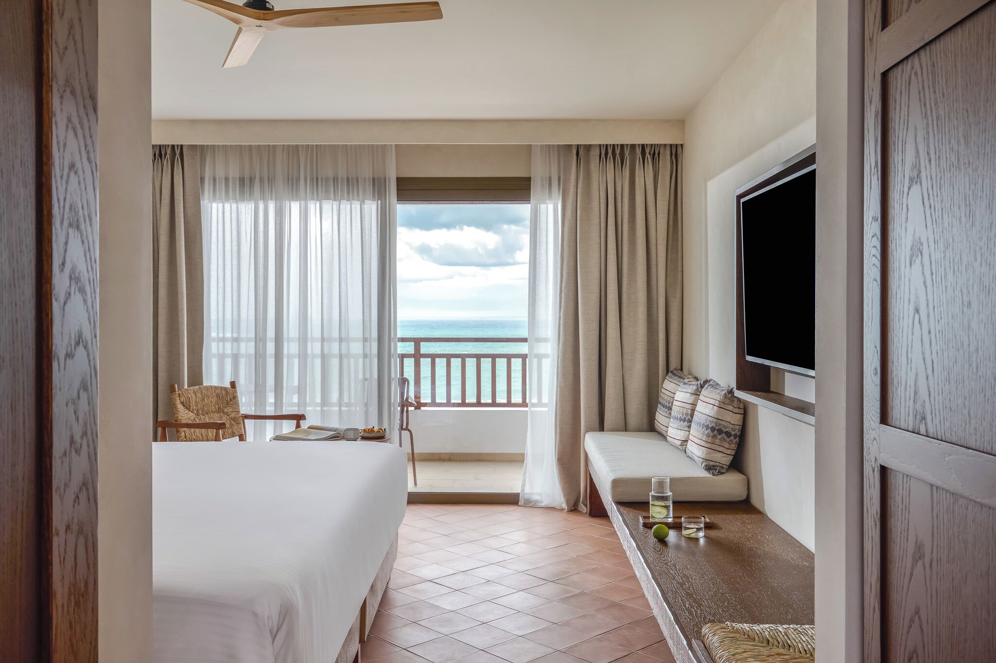 Quarto de hotel com cama, área de estar com almofadas, TV e varanda com vista para o mar.