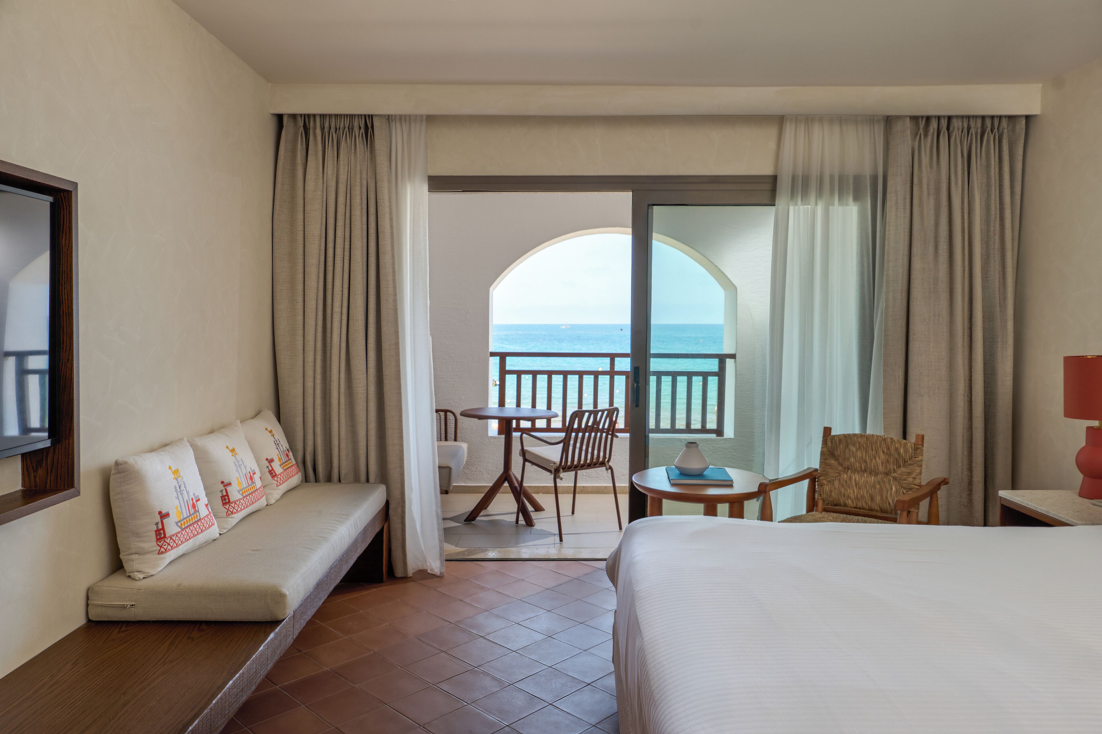 Une chambre d’hôtel avec un lit, un canapé agrémenté de coussins décoratifs, une petite table avec une lampe, une chaise et un balcon offrant une vue sur la mer.