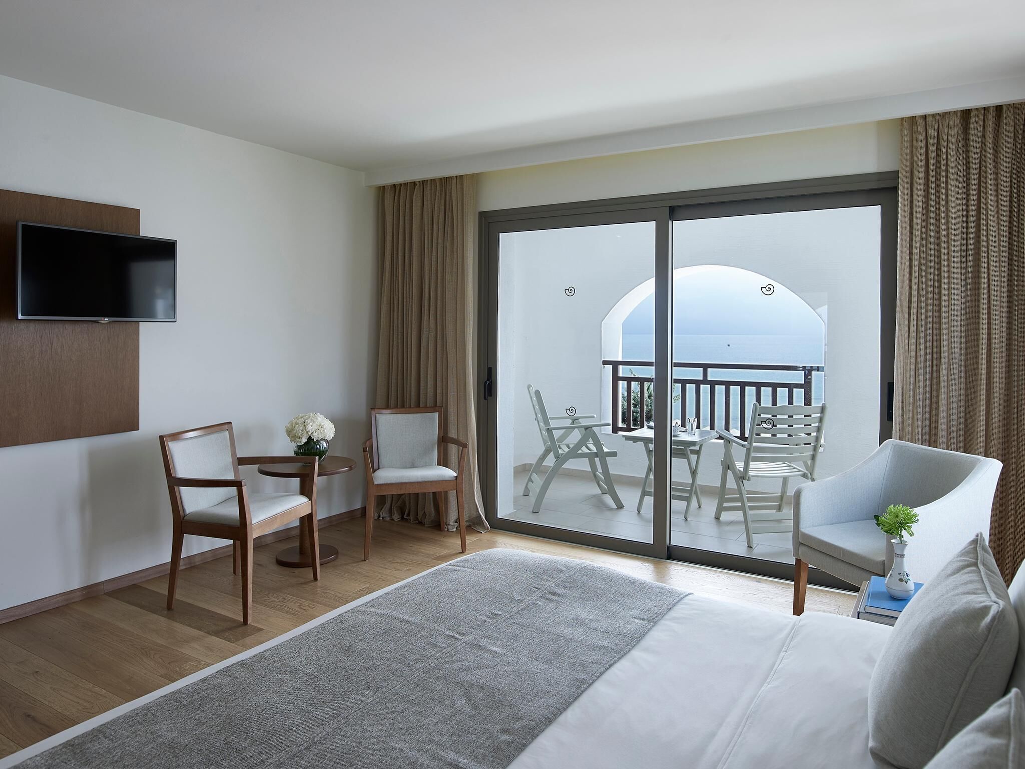 A hotel room with a bed, a TV, a balcony with chairs, and a view of the sea.