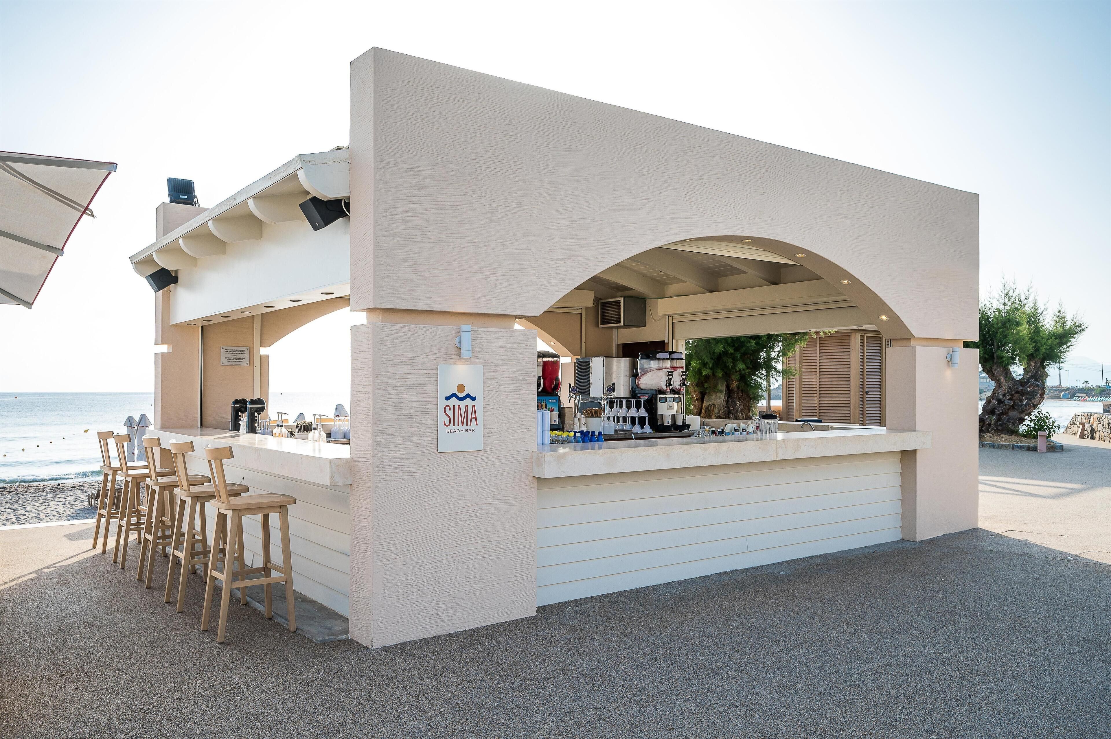 A beach bar with a white exterior, bar stools, and a view of the ocean.