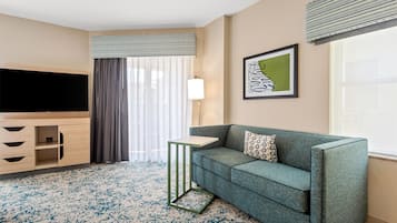 A modern hotel room with a grey sofa, a small table, a TV, and a window with curtains.