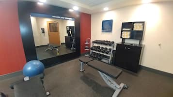 Un gimnasio con equipo de ejercicio, una banca, un espejo adosado a la pared y un estante con mancuernas.