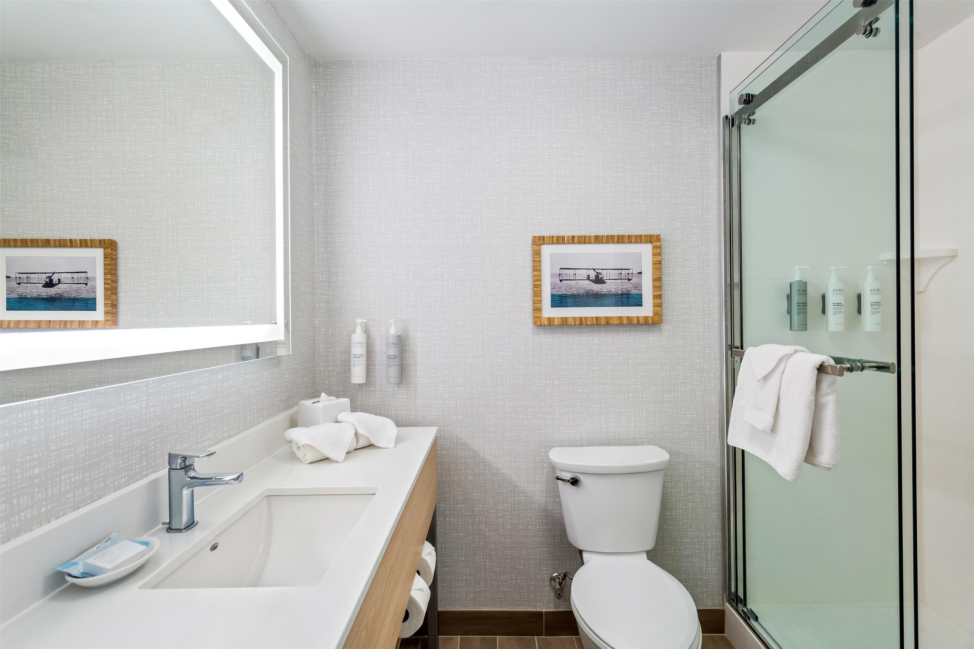 Un baño moderno con lavamanos, espejo y retrete blancos, cuadros en la pared y una cabina de ducha de vidrio.