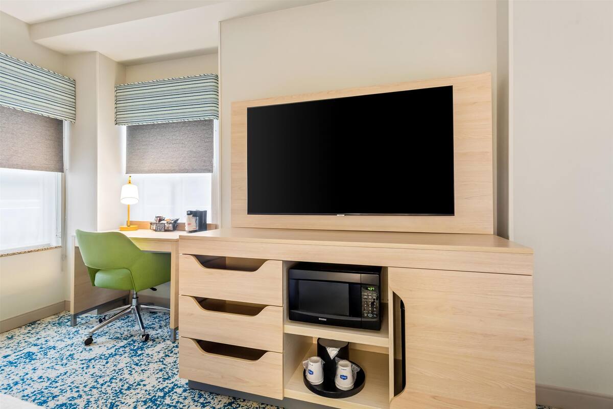 Habitación de hotel con un escritorio de madera, una silla verde, un televisor de pantalla plana, un microondas y una cafetera.