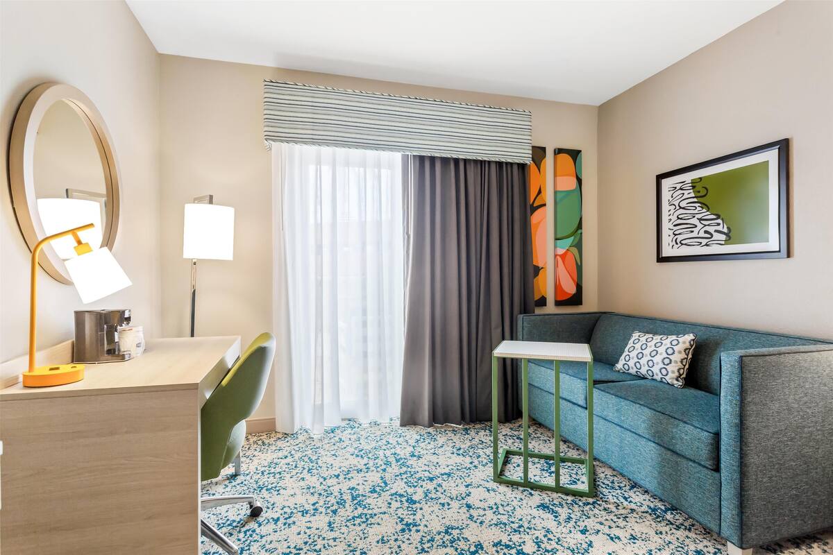 Una habitación de hotel con escritorio, silla, sofá y un ventanal con cortinas.