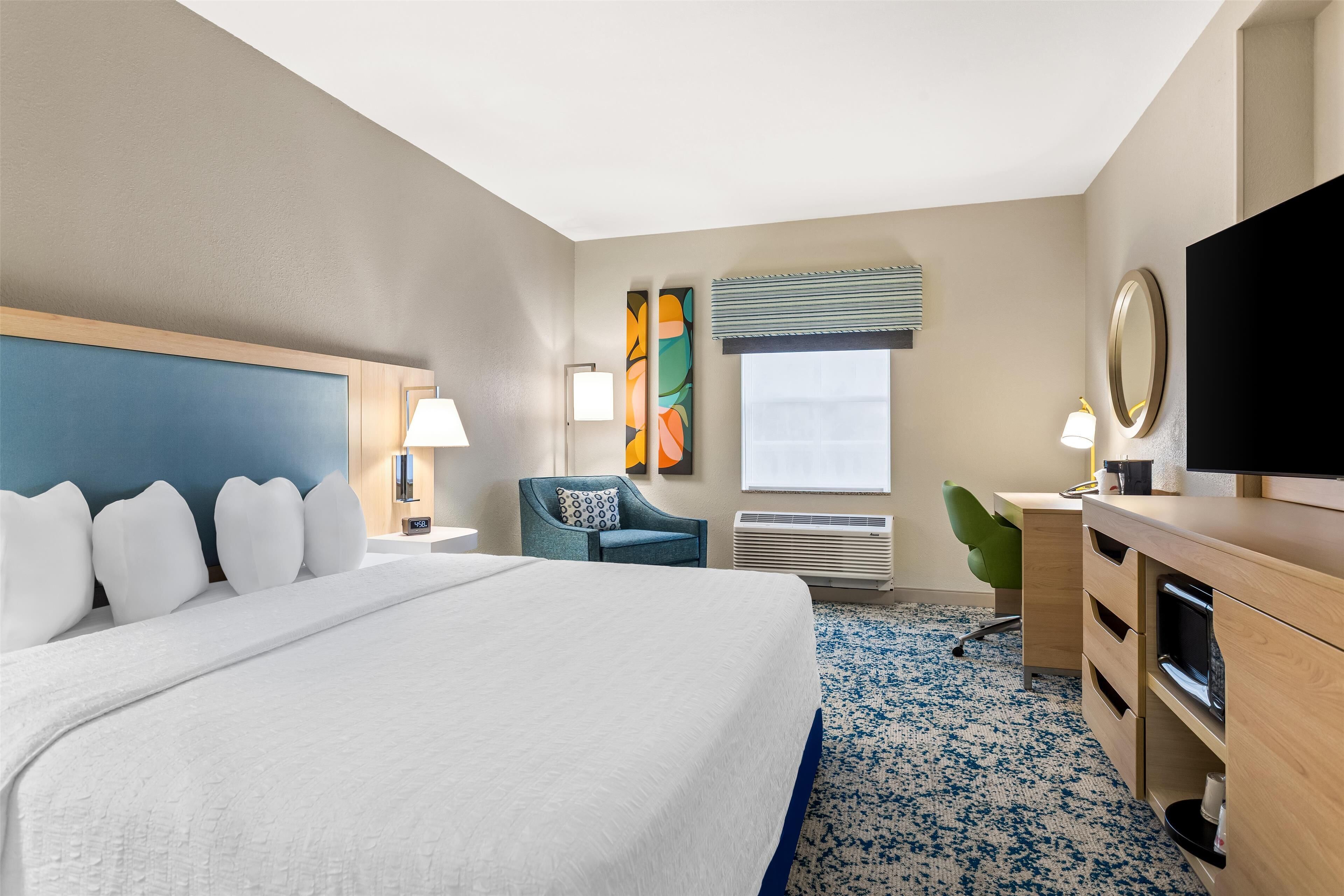 A hotel room with a large bed, a desk with a flat-screen TV, a chair, a desk lamp, and a window with blinds.