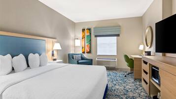 A hotel room with a large bed, a desk with a flat-screen TV, a chair, a desk lamp, and a window with blinds.