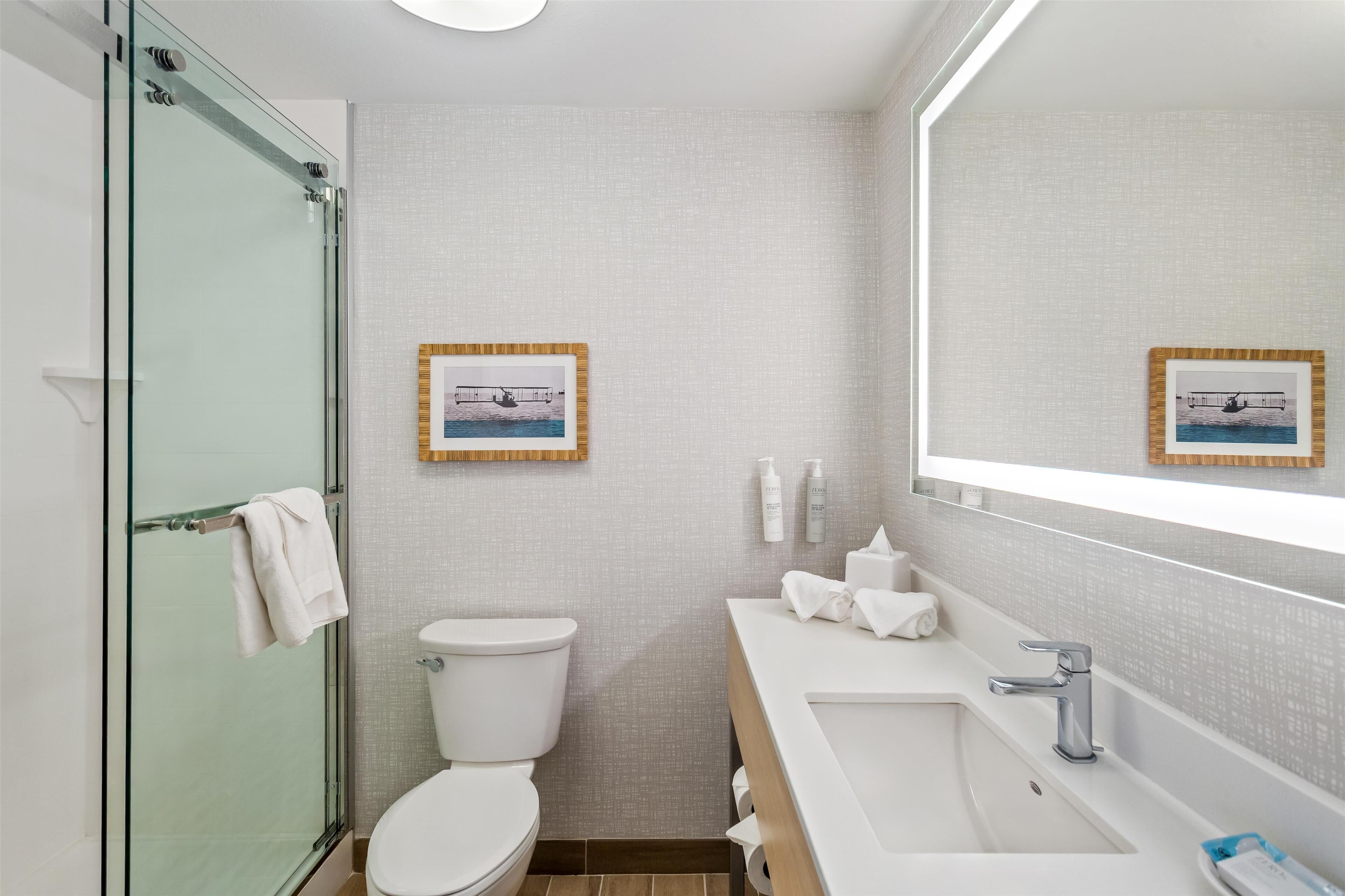 Un baño moderno con cabina de ducha de vidrio, inodoro blanco, lavabo con espejo y cuadros enmarcados en la pared.