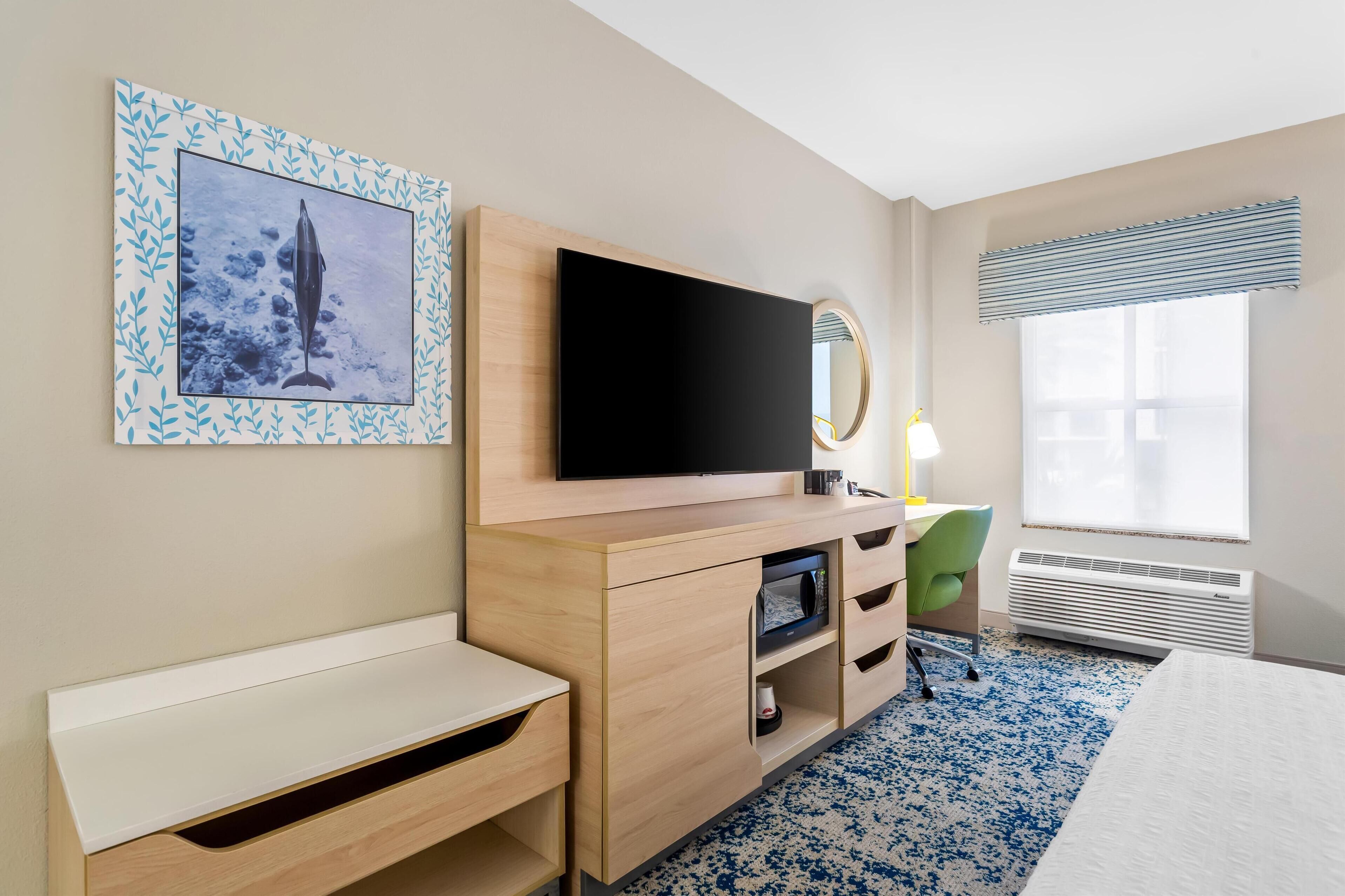 A hotel room with a flat-screen TV, a desk, a chair, a bed, a window with blinds, and a framed picture on the wall.