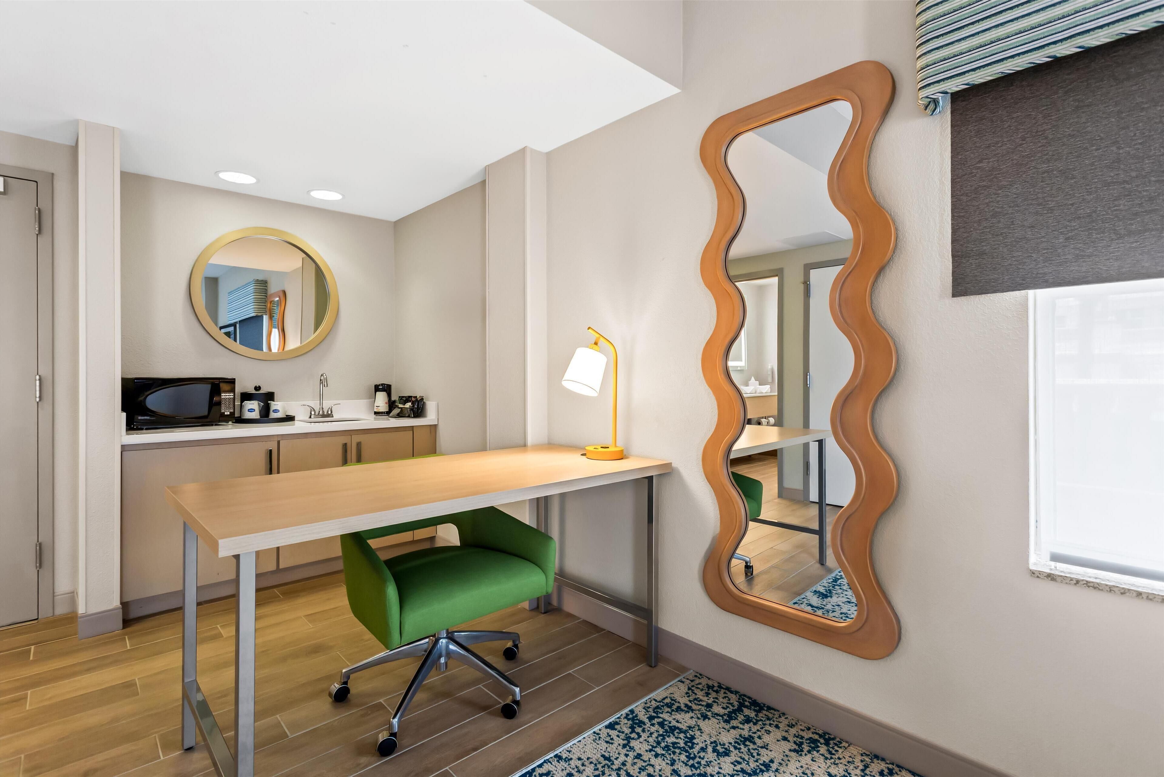 A modern room with a wooden desk, a green chair, a round mirror, and a wavy mirror on the wall.