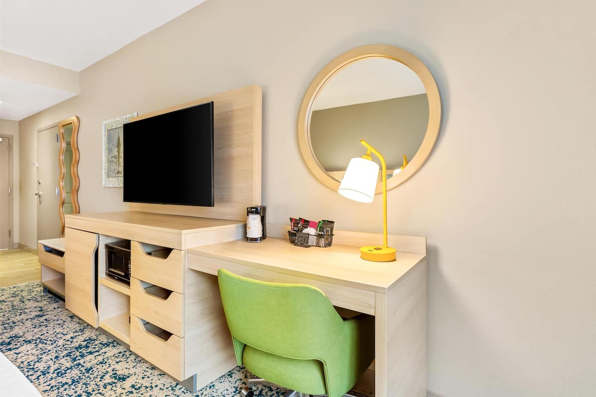 Habitación de hotel con un escritorio de madera, una silla verde, un televisor, un espejo y una lámpara.