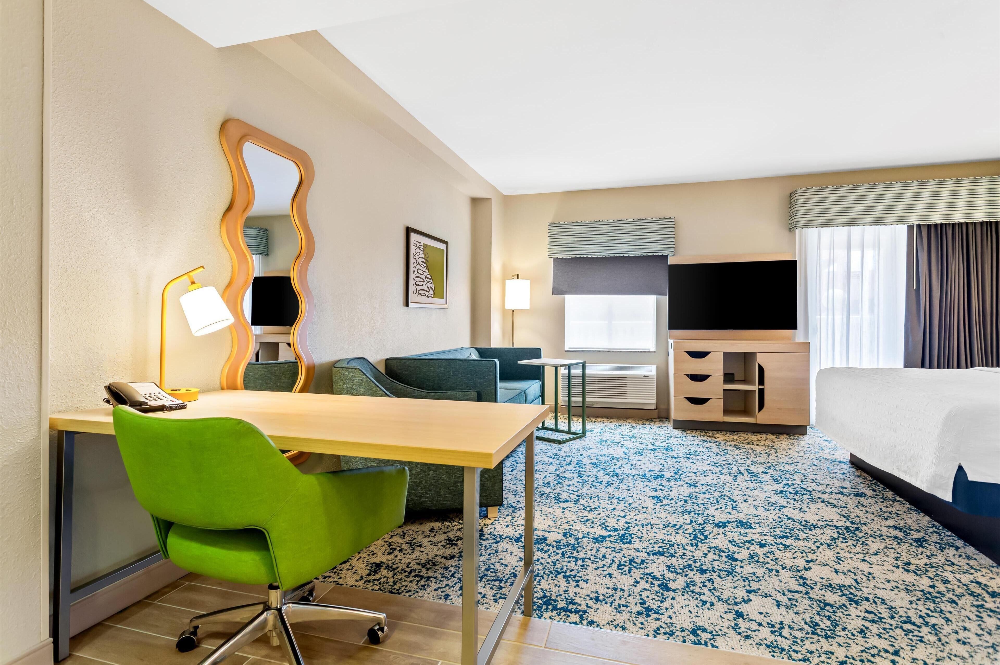 Habitación de hotel con cama, escritorio con silla, televisor, lámpara, espejo y ventana con cortinas.