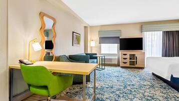 Habitación de hotel con cama, escritorio con silla, televisor, lámpara, espejo y ventana con cortinas.