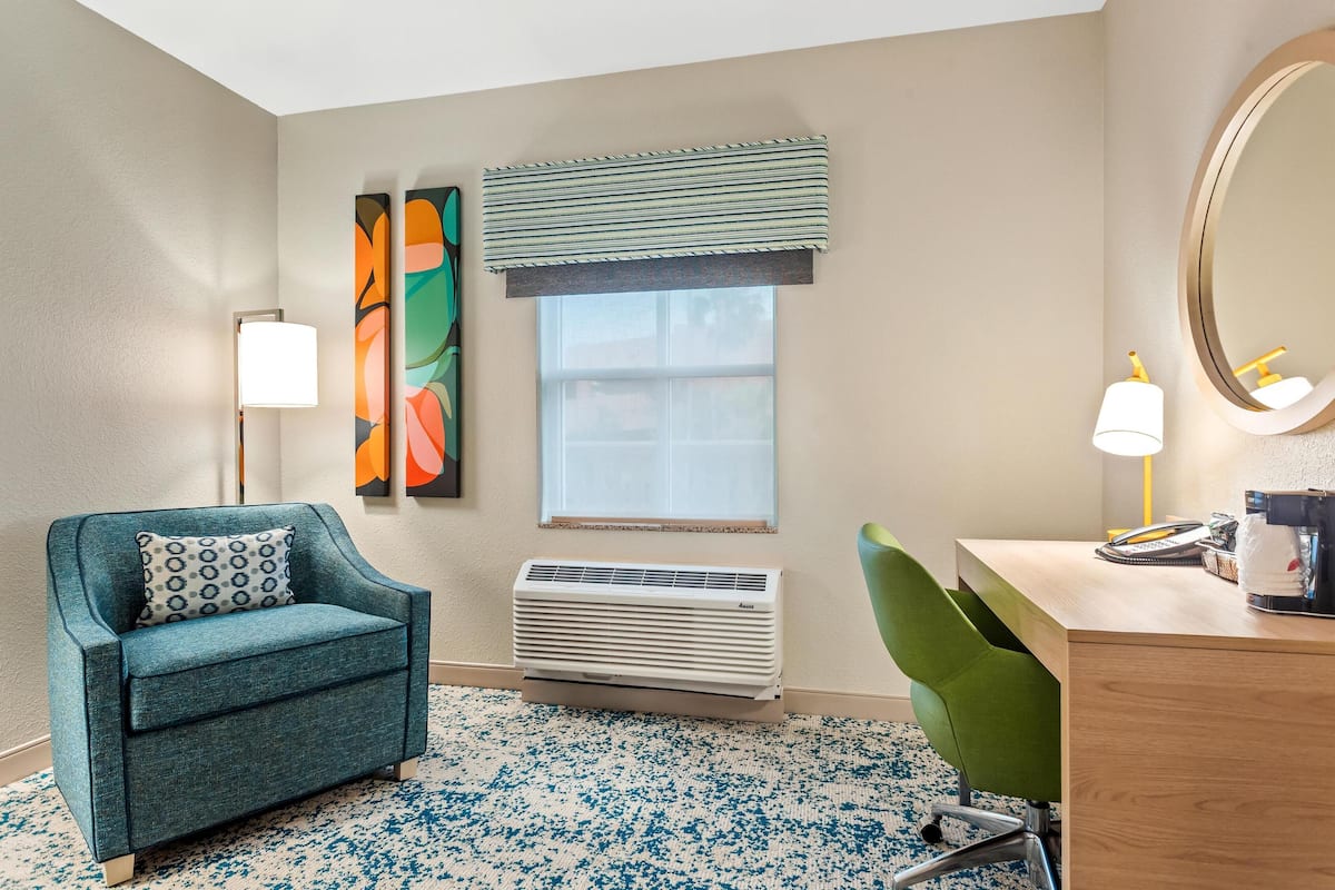 Una habitación con un sillón azul verdoso, un escritorio con lámpara, un espejo grande, una ventana con persianas y un aire acondicionado instalado en la pared.