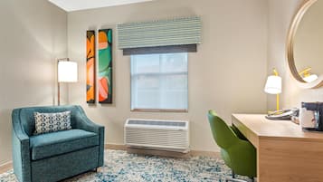 A room with a teal armchair, a desk with a lamp, a large mirror, a window with blinds, and a wall-mounted air conditioner.