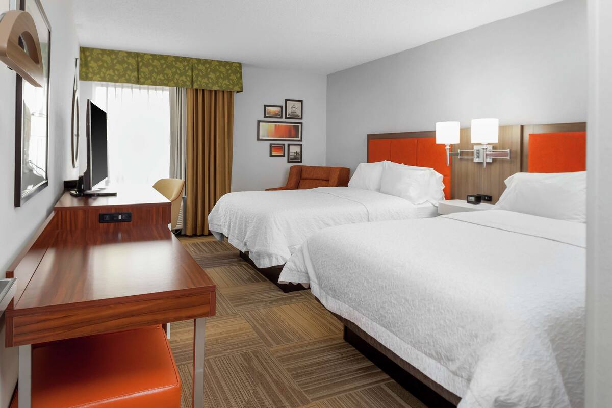 A hotel room with two beds, a desk, a TV, and a window with curtains.