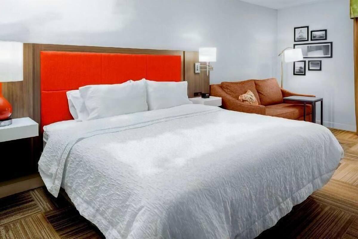 A hotel room with a large bed, a red headboard, a brown sofa, a nightstand, a lamp, and framed pictures on the wall.