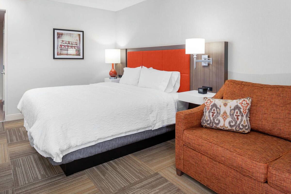 A hotel room with a large bed, a sofa, a nightstand with a lamp, and a framed picture on the wall.