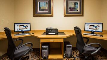 A hotel room with a desk, two computers, two chairs, and two framed pictures on the wall.