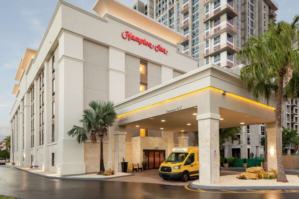 A Hampton Inn hotel entrance with a clear sign, a welcoming bench, and a yellow shuttle service.