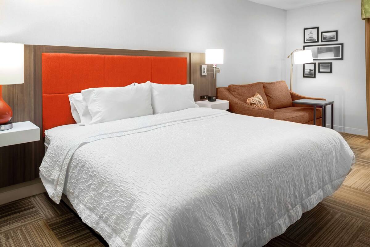 A hotel room with a large bed, a nightstand, a lamp, a sofa, and framed pictures on the wall.