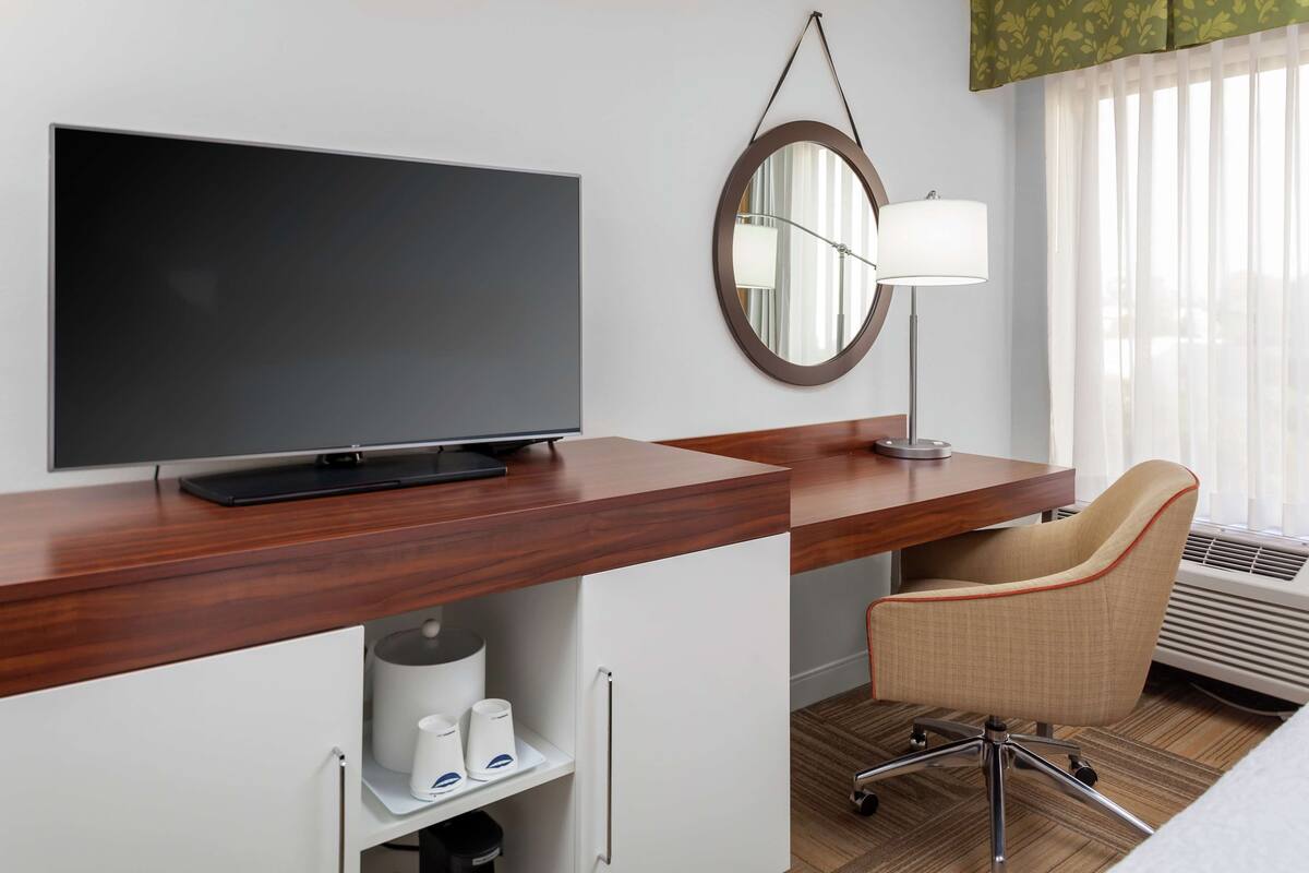 A hotel room with a flat-screen TV on a wooden desk, a round mirror, a lamp, a chair, and a bed.