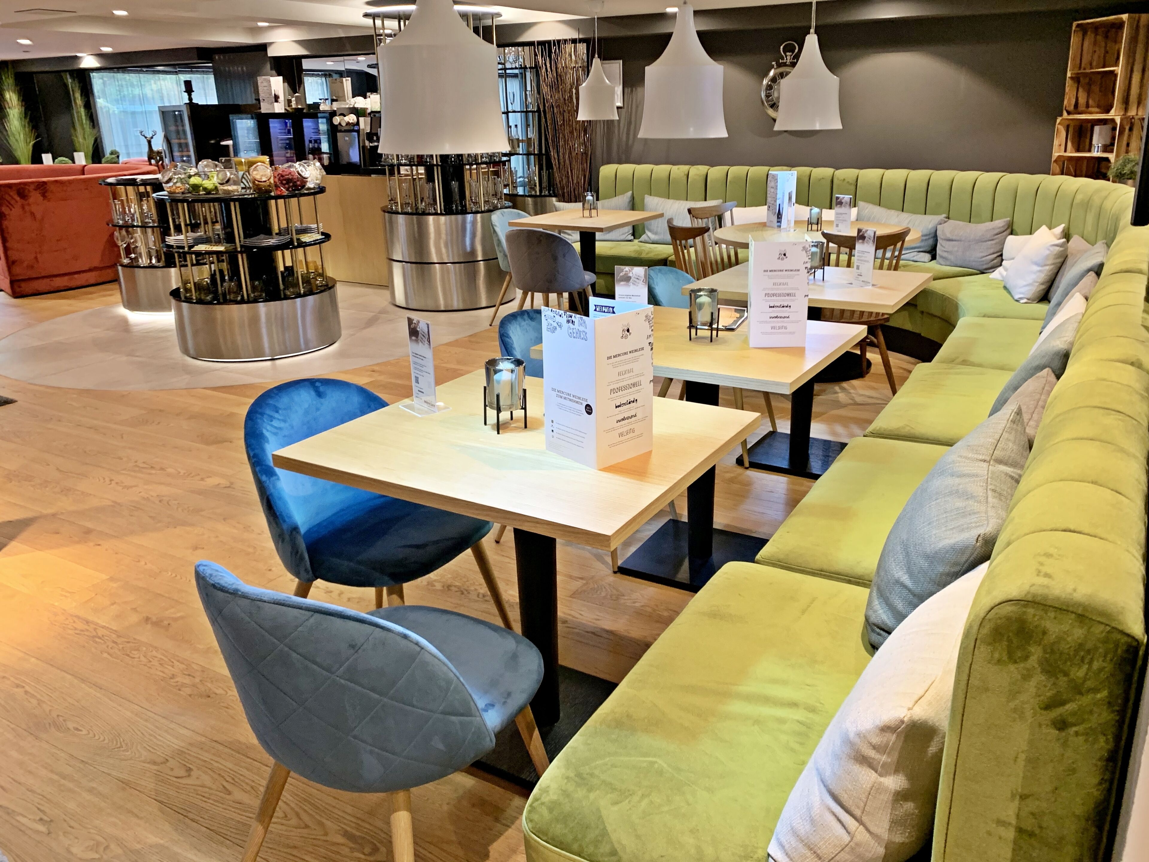 A modern restaurant with green upholstered seating, wooden tables, and hanging pendant lights.