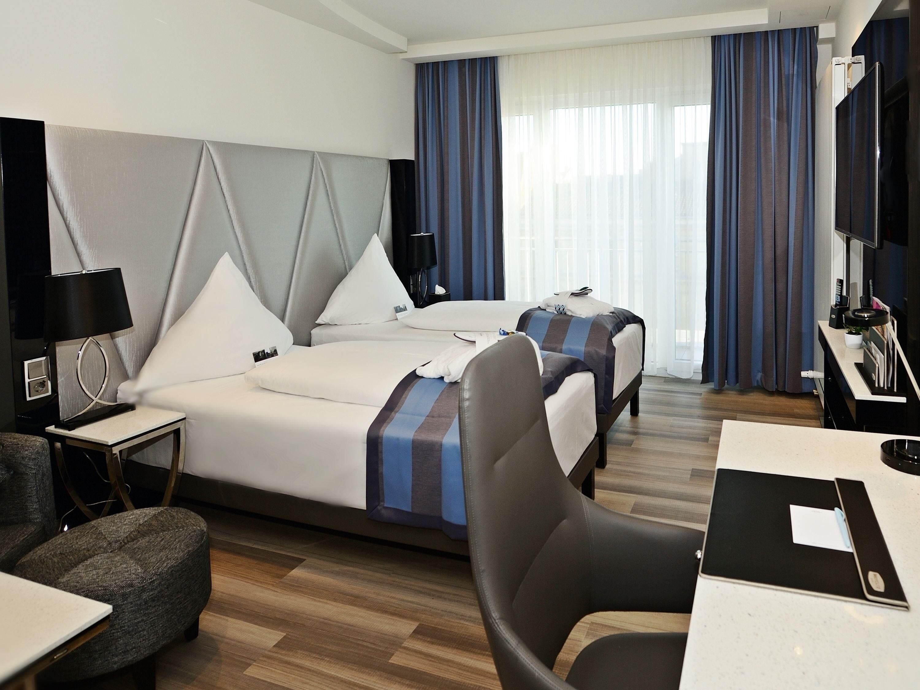 A hotel room with two beds, a desk, a chair, a TV, and a window with curtains.