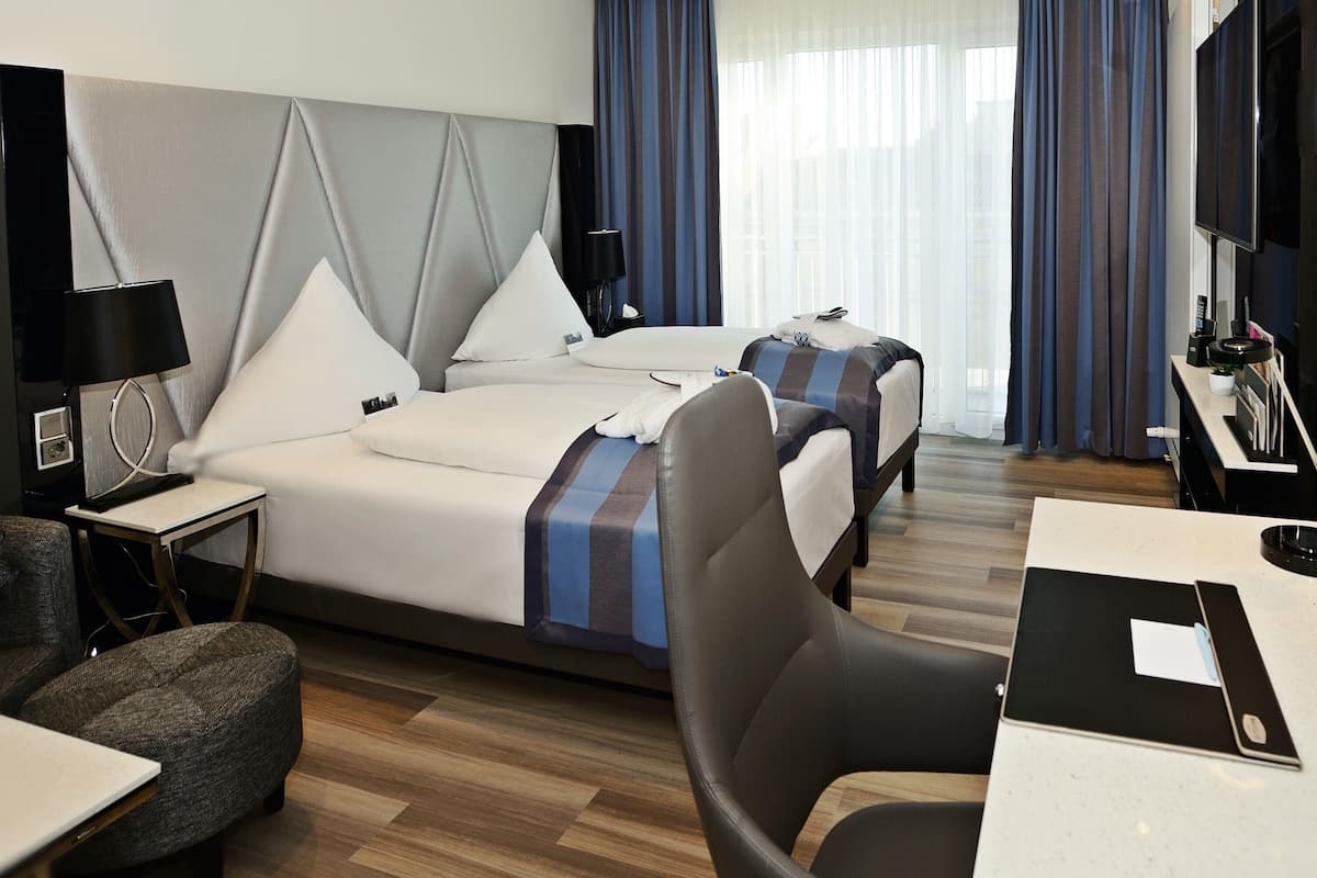 A hotel room with two beds, a desk, a chair, a TV, and a window with curtains.