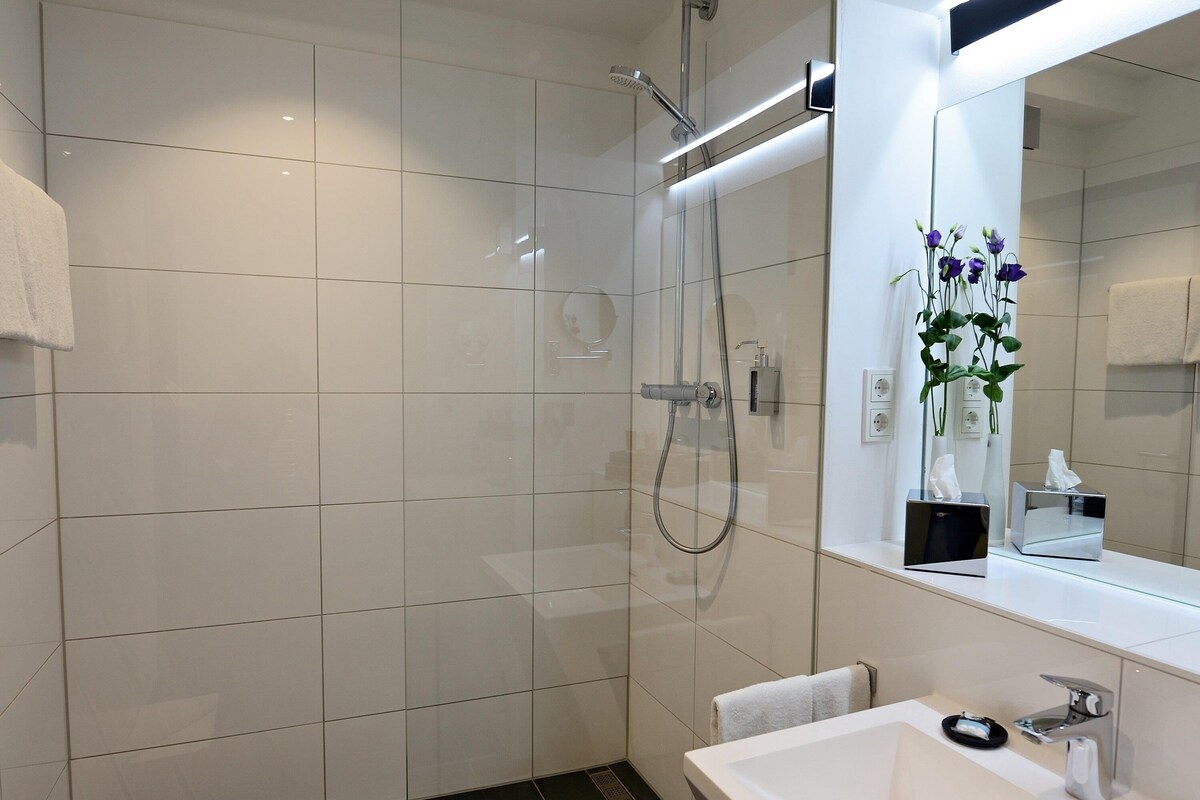 Un baño moderno con ducha envuelta en vidrio, un lavabo blanco y un espejo con un florero.
