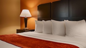 A hotel room with a bed, white pillows, a red throw blanket, a nightstand with a lamp, and a dark headboard. - Best Western Inn & Suites - Lake Charles (Lake Charles)