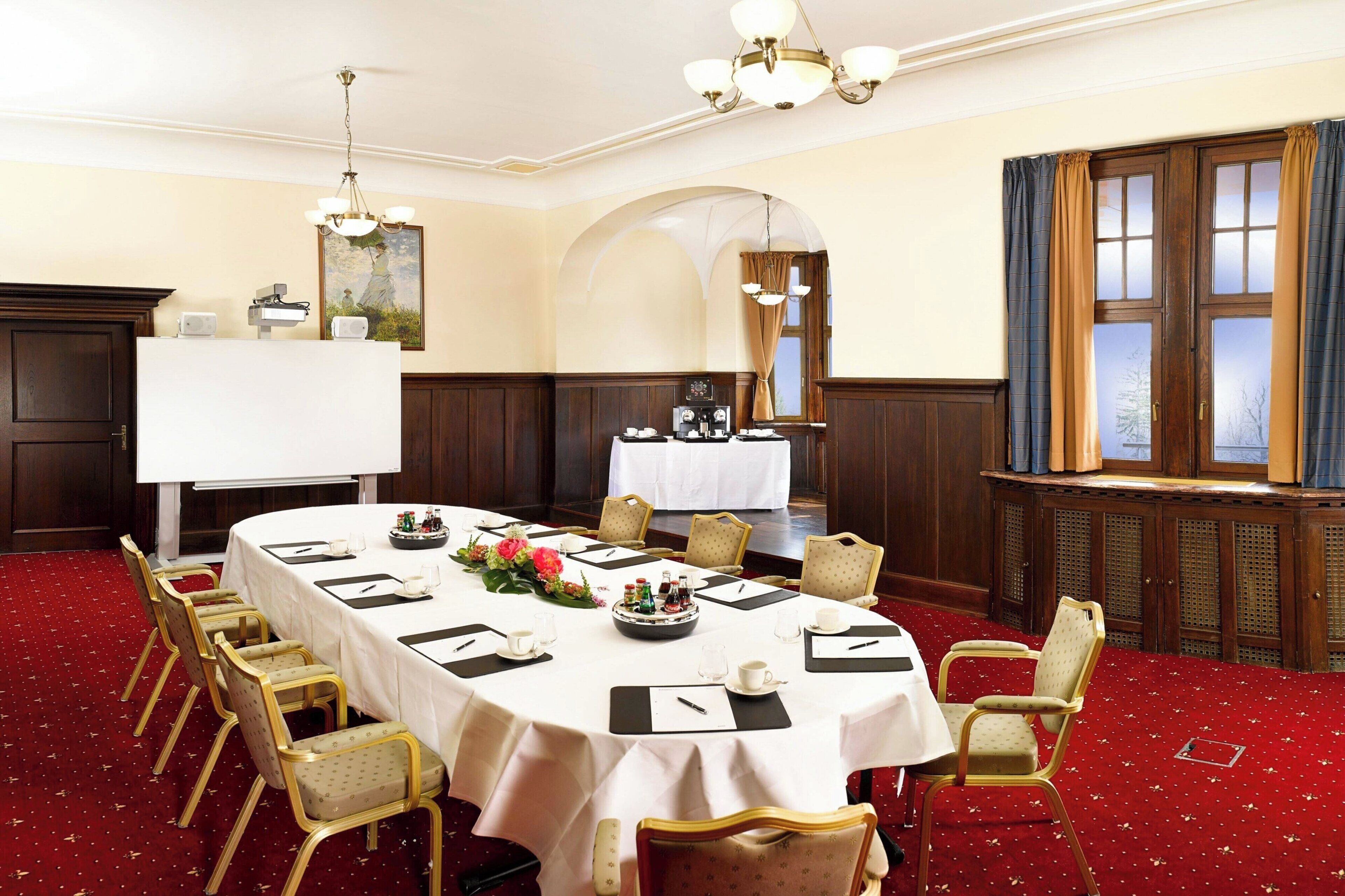 A conference room with a long table set for a meeting, surrounded by chairs, with a chandelier and a window with curtains.