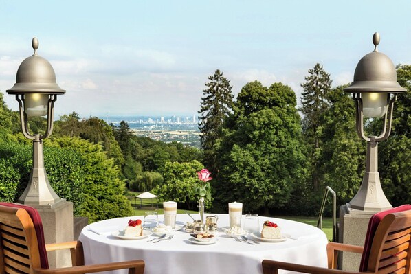 Mesa redonda preparada para una comida, con vistas a un paisaje urbano, rodeada de árboles y dos faroles decorativos.