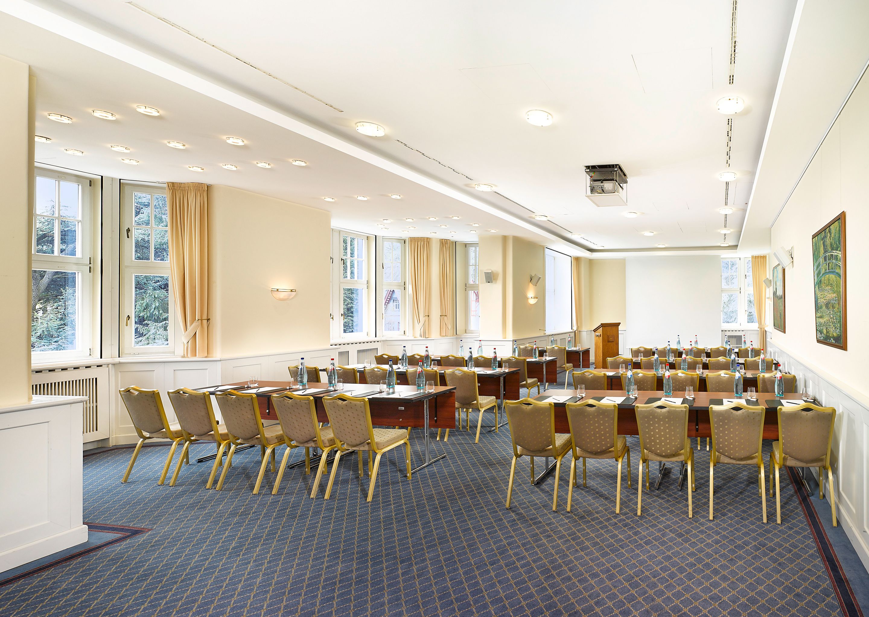 A conference room with multiple tables and chairs, large windows with curtains, and a blue patterned carpet.
