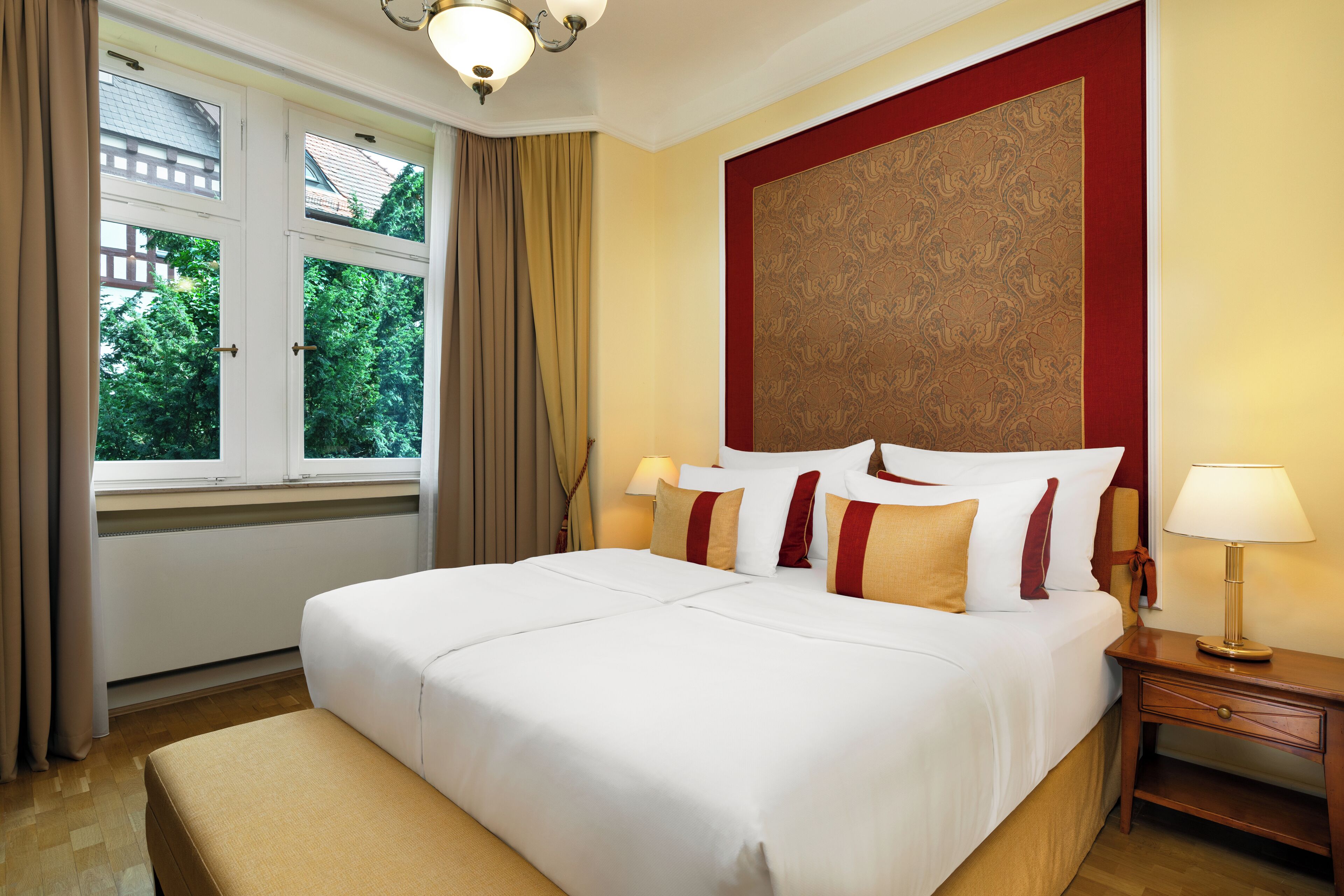 A hotel room with a large bed, a nightstand with a lamp, a window with curtains, and a decorative wall panel.